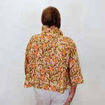 A woman viewed from behind wearing a short-sleeved collared blouse with an orange, green, and brown floral pattern on a beige background, paired with white pants, set against a plain white wall.