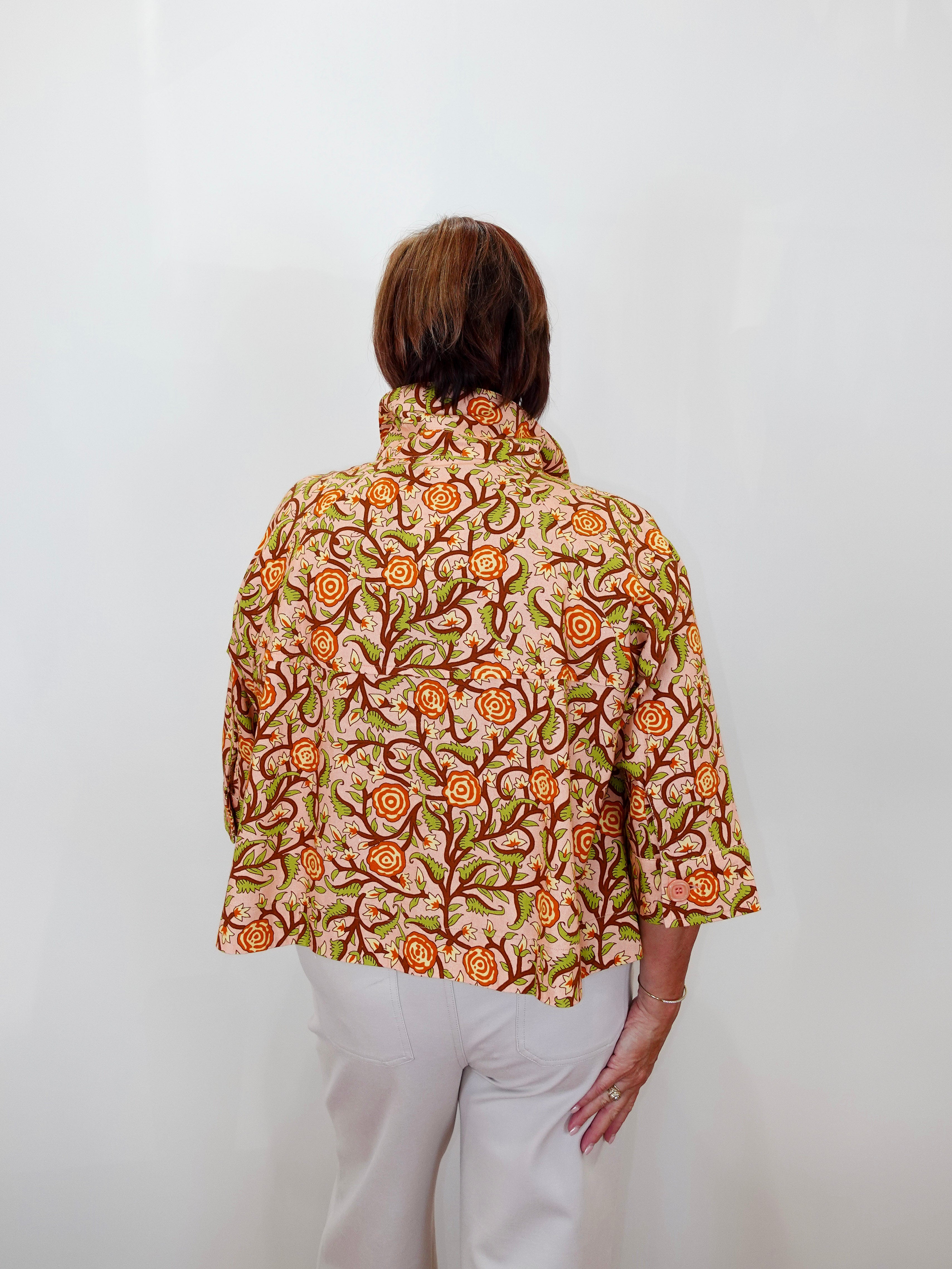 A woman viewed from behind wearing a short-sleeved collared blouse with an orange, green, and brown floral pattern on a beige background, paired with white pants, set against a plain white wall.