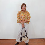 A woman stands against a white wall wearing a yellow and orange patterned blouse, white wide-leg pants, and black heeled sandals, while holding a beige and black handbag.