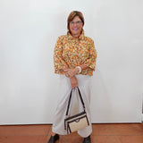A woman stands against a white wall wearing a yellow and orange patterned blouse, white wide-leg pants, and black heeled sandals, while holding a beige and black handbag.