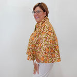 A woman with glasses is shown from the side, wearing a beige short-sleeve blouse with a vibrant orange and green floral pattern, paired with white pants, against a plain white wall.
