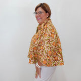 A woman with glasses is shown from the side, wearing a beige short-sleeve blouse with a vibrant orange and green floral pattern, paired with white pants, against a plain white wall.