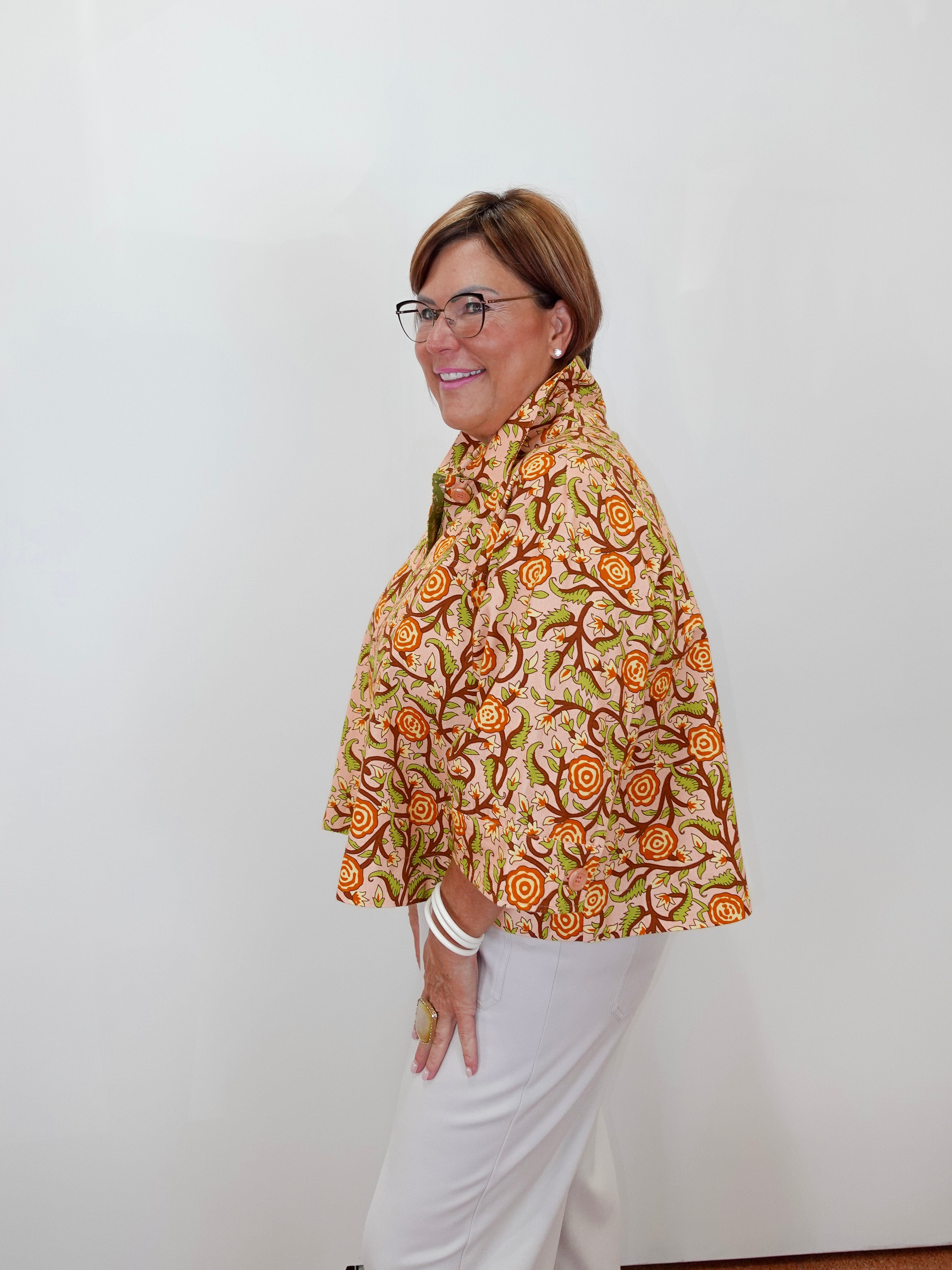 A woman with glasses is shown from the side, wearing a beige short-sleeve blouse with a vibrant orange and green floral pattern, paired with white pants, against a plain white wall.