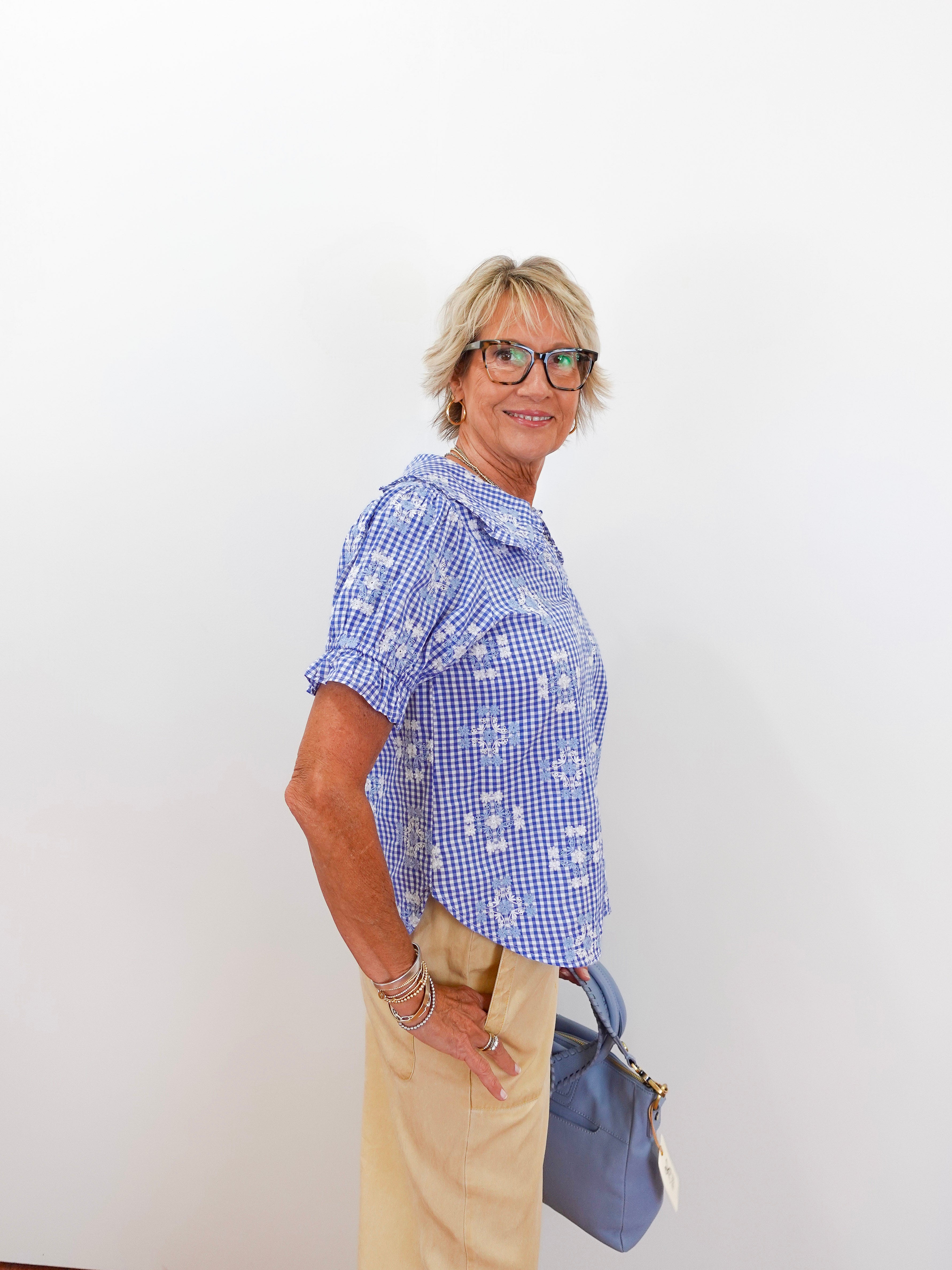 Woman wearing a blue checkered shirt and beige pants, holding a blue handbag against a white background