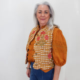 An older woman with gray hair stands against a plain white wall wearing a distinctive orange and white checkered embroidered blouse with puff sleeves and blue jeans.