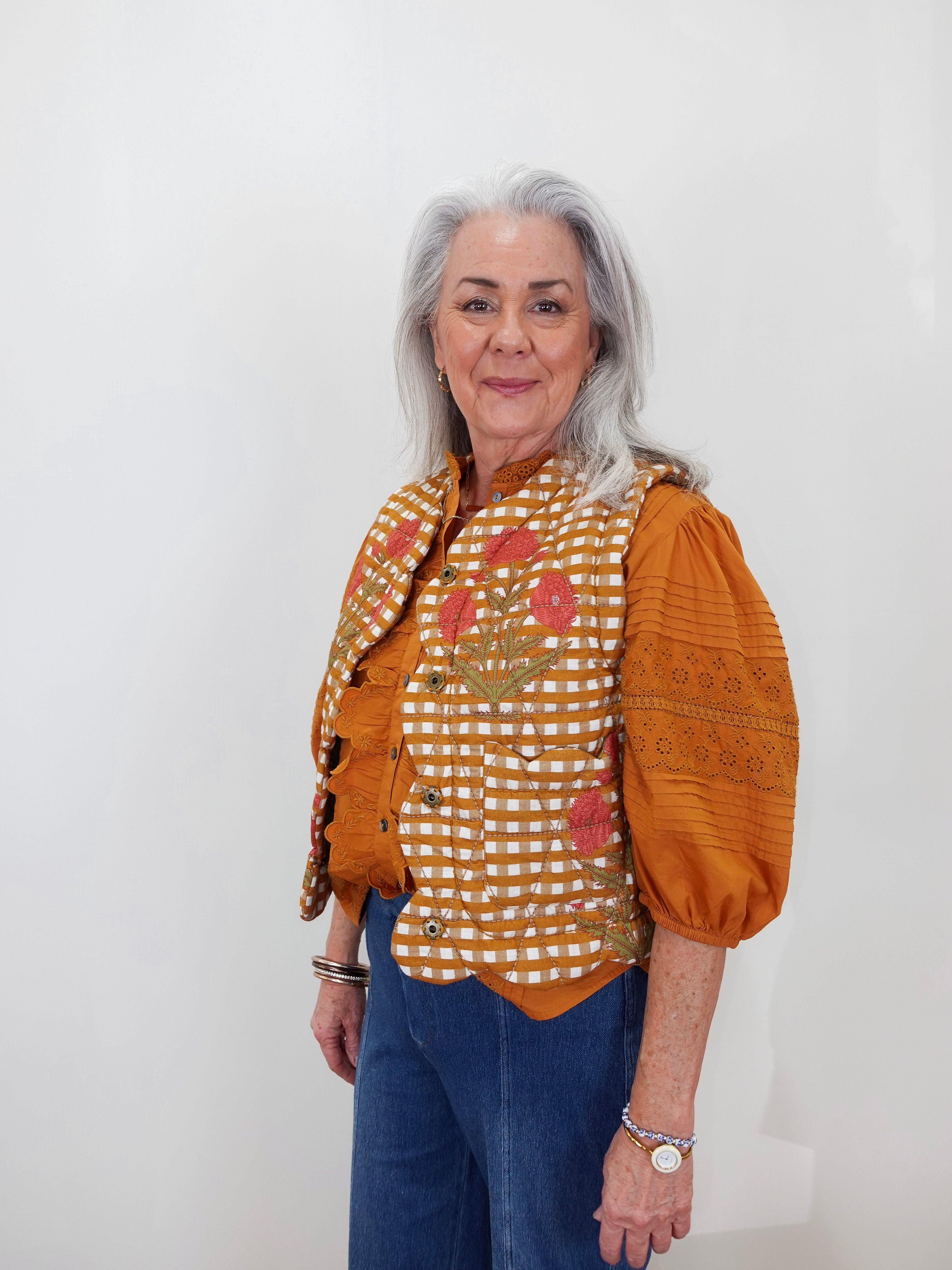 An older woman with gray hair stands against a plain white wall wearing a distinctive orange and white checkered embroidered blouse with puff sleeves and blue jeans.