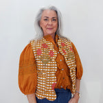 An older woman with gray hair is wearing a rust-orange blouse with puffed sleeves, a white and orange gingham vest with pink floral embroidery, and blue jeans, standing against a plain white wall.