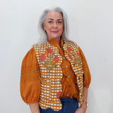An older woman with gray hair is wearing a rust-orange blouse with puffed sleeves, a white and orange gingham vest with pink floral embroidery, and blue jeans, standing against a plain white wall.