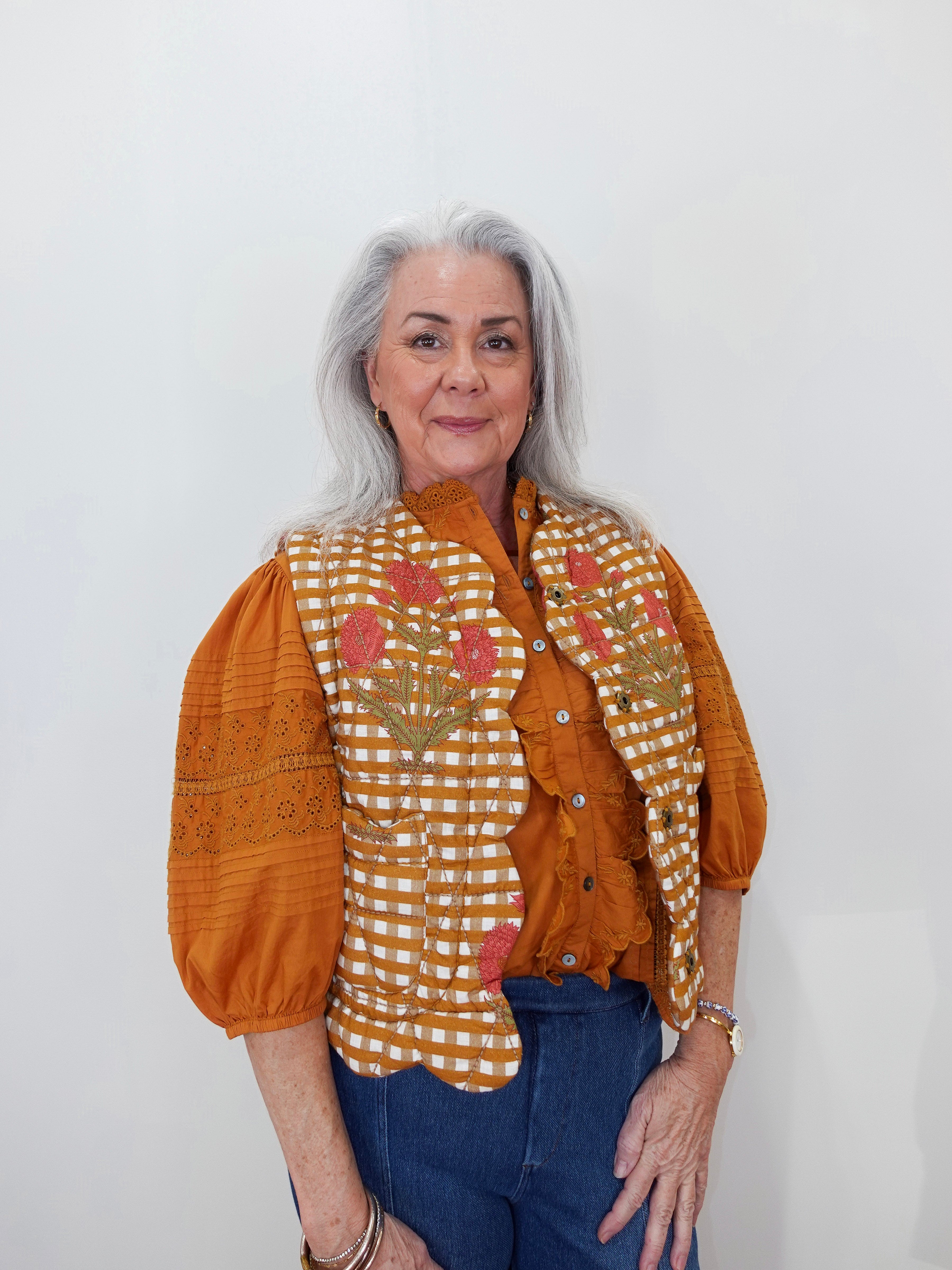 An older woman with gray hair is wearing a rust-orange blouse with puffed sleeves, a white and orange gingham vest with pink floral embroidery, and blue jeans, standing against a plain white wall.
