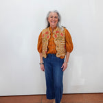 An older woman with gray hair stands against a plain white wall wearing a rust-orange blouse, a white vest with orange and red floral embroidery, blue cropped jeans, and beige platform sandals.
