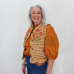 An older woman with gray hair stands against a plain white wall wearing a distinctive orange and white gingham vest with floral embroidery, orange puff sleeves, and blue jeans.