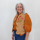 An older woman with gray hair stands against a plain white wall wearing a distinctive orange and white gingham vest with floral embroidery, orange puff sleeves, and blue jeans.