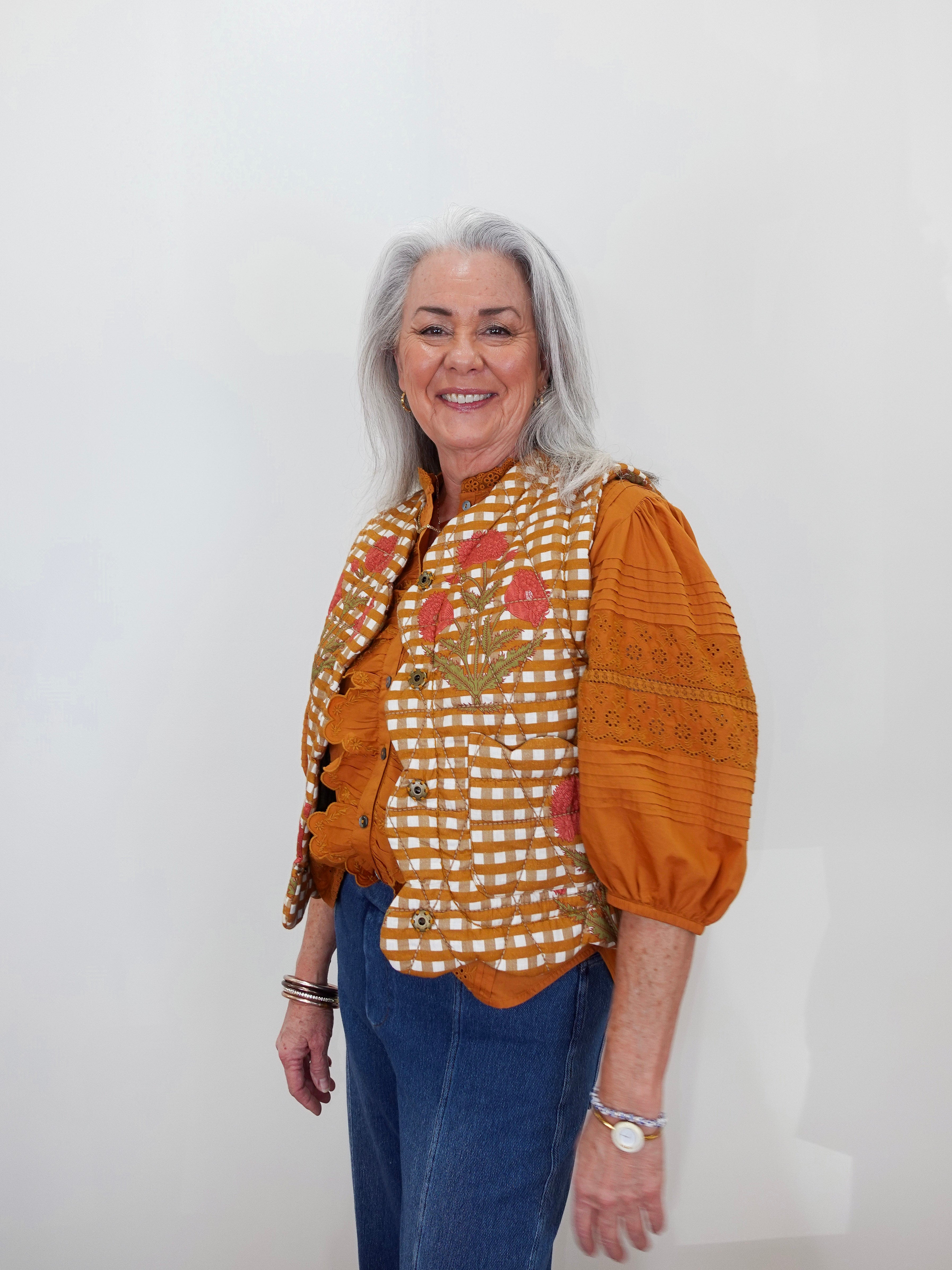 An older woman with gray hair stands against a plain white wall wearing a distinctive orange and white gingham vest with floral embroidery, orange puff sleeves, and blue jeans.