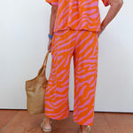 Person wearing an orange and pink zebra print outfit with a woven bag against a white wall.