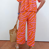Person wearing an orange and pink zebra print outfit with a woven bag against a white wall.