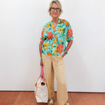 Woman wearing a colorful floral blouse and beige pants, holding a white bag against a white wall.