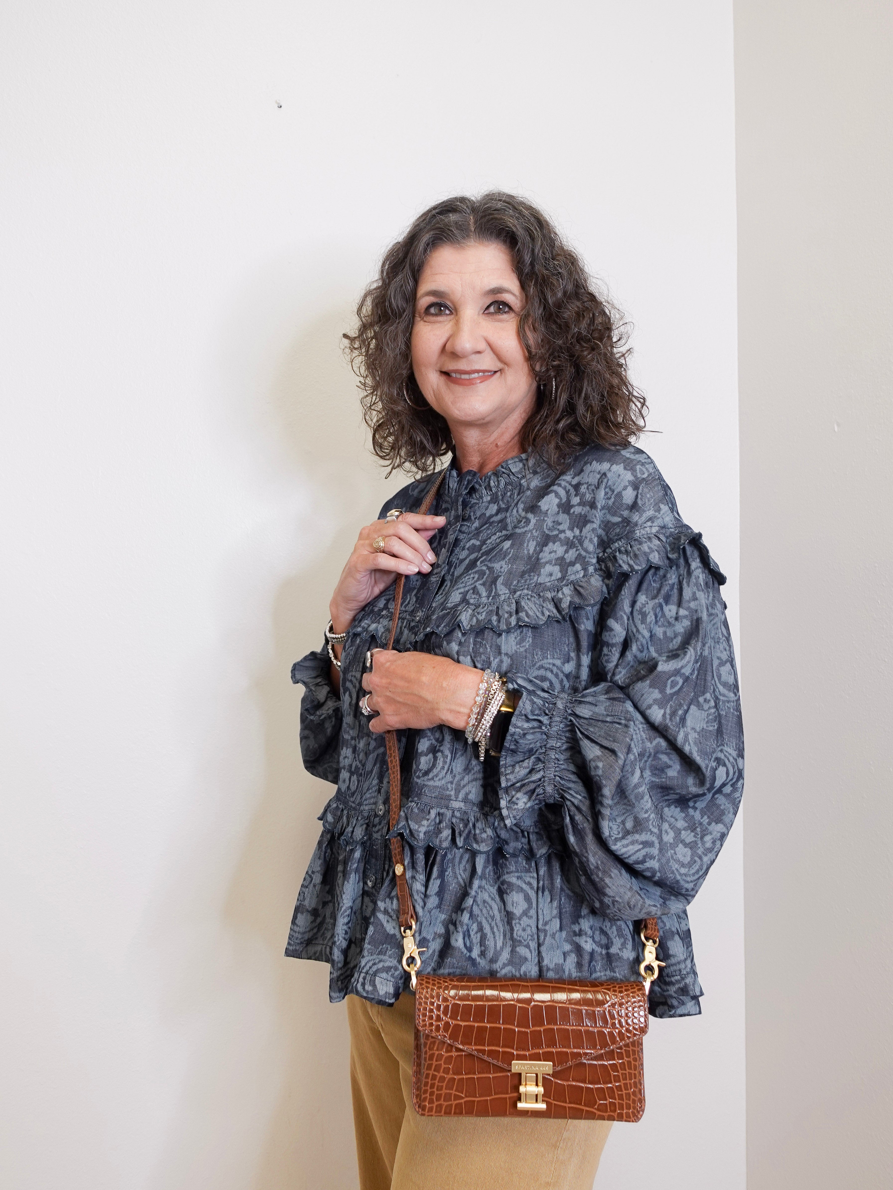Woman wearing a patterned blouse and holding a brown handbag against a white background