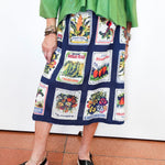 A person wearing a navy midi skirt with a vintage vegetable patchwork print and a bright green top, paired with black wedge sandals, standing against a white wall.