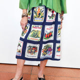 A person wearing a navy midi skirt with a vintage vegetable patchwork print and a bright green top, paired with black wedge sandals, standing against a white wall.