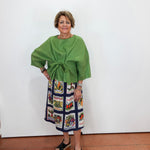 A woman stands against a plain white wall wearing a bright green cropped top with a gathered waist and a navy midi skirt featuring colorful vintage-style illustrated panels. She is wearing black slide sandals.