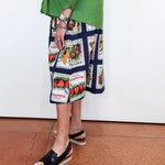 A person wearing a green top and a navy midi skirt with a vegetable patchwork print, paired with black slide sandals, standing against a white wall on a terracotta tiled floor.
