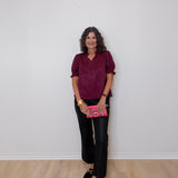 Woman wearing a burgundy top and black pants holding a pink book against a white wall.