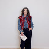 Woman wearing a denim jacket over a red patterned shirt with black pants and white sneakers, holding a white handbag against a plain background.