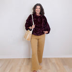 Woman wearing a dark patterned top, beige pants, and tan shoes, holding a beige handbag against a white wall.