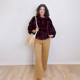 Woman wearing a dark patterned top, beige pants, and tan shoes, holding a beige handbag against a white wall.