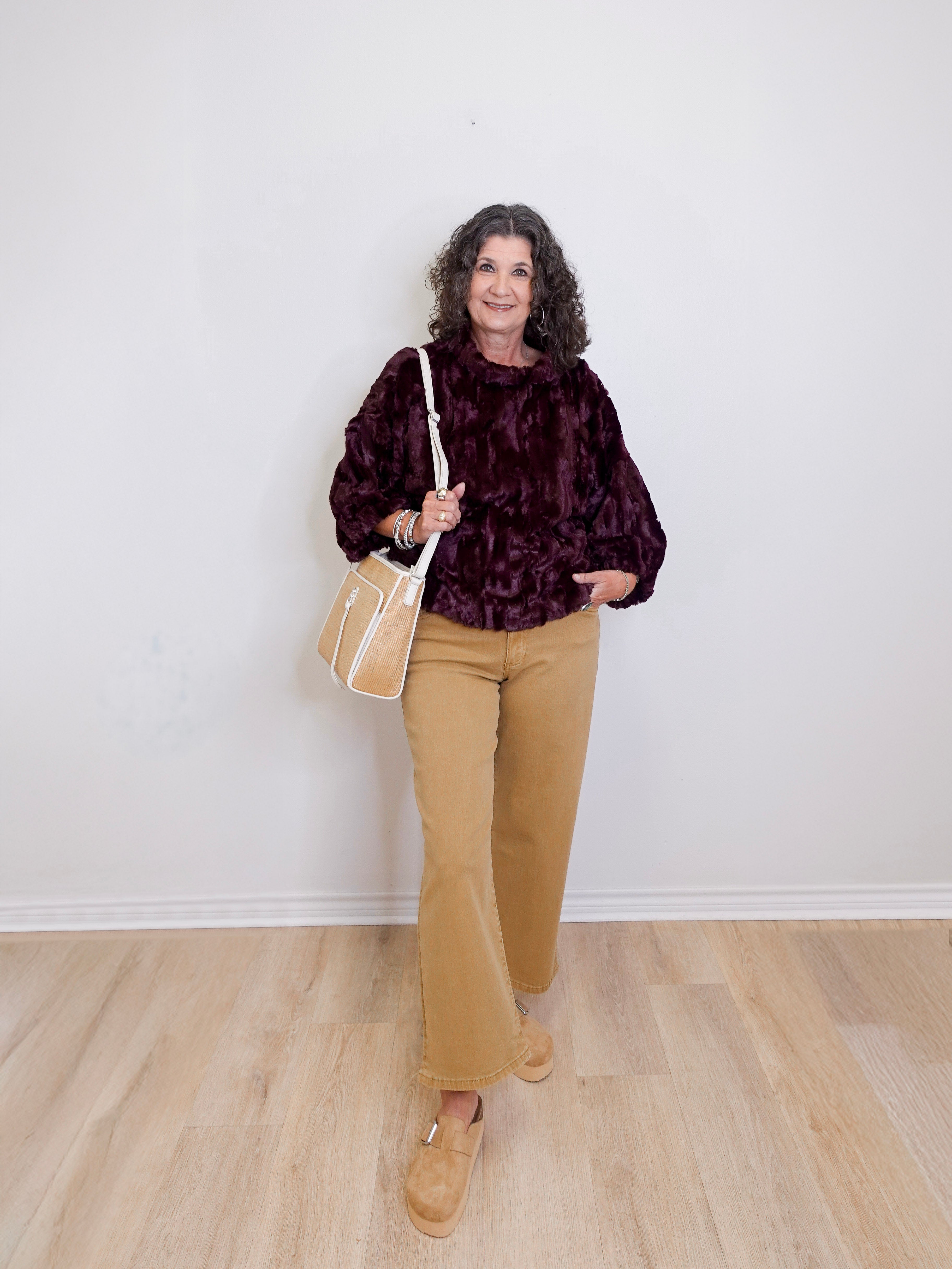 Woman wearing a dark patterned top, beige pants, and tan shoes, holding a beige handbag against a white wall.
