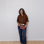 Woman wearing a brown shirt, blue jeans, and a patterned clutch against a white wall.