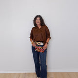 Woman wearing a brown shirt, blue jeans, and a patterned clutch against a white wall.