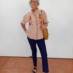Woman wearing a beige embroidered blouse, blue pants, and black sandals against a white wall.