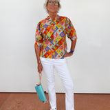 Woman wearing a colorful patterned shirt and white pants, holding a blue handbag against a plain background.