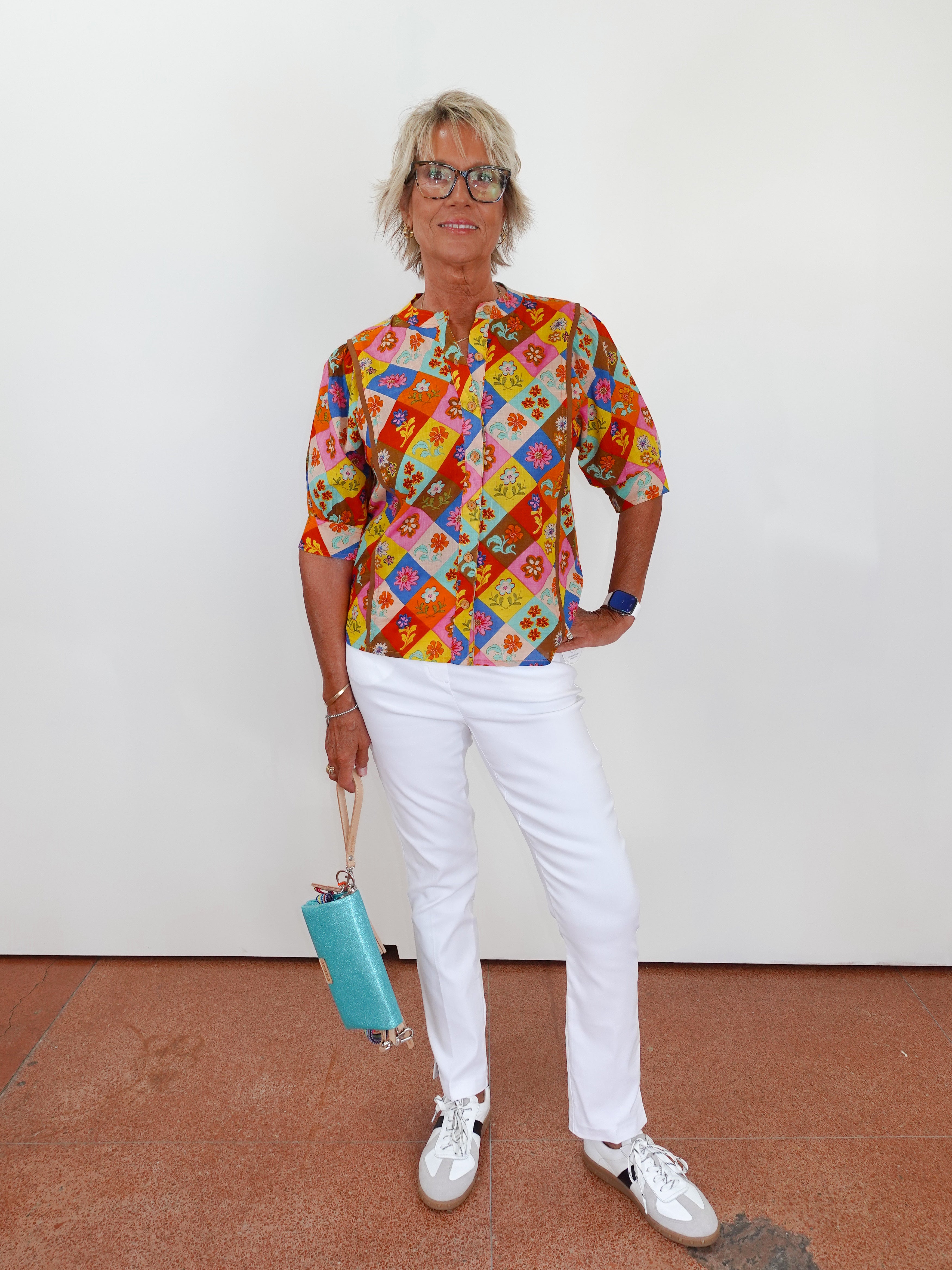 Woman wearing a colorful patterned shirt and white pants, holding a blue handbag against a plain background.