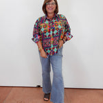 A woman modeling a colorful, patterned long-sleeve button-up shirt with light blue flared jeans and dark sandals, standing against a plain white wall.