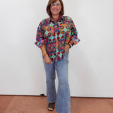 A woman modeling a colorful, patterned long-sleeve button-up shirt with light blue flared jeans and dark sandals, standing against a plain white wall.