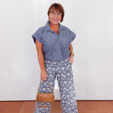 Woman wearing a blue shirt and patterned pants standing against a white wall.