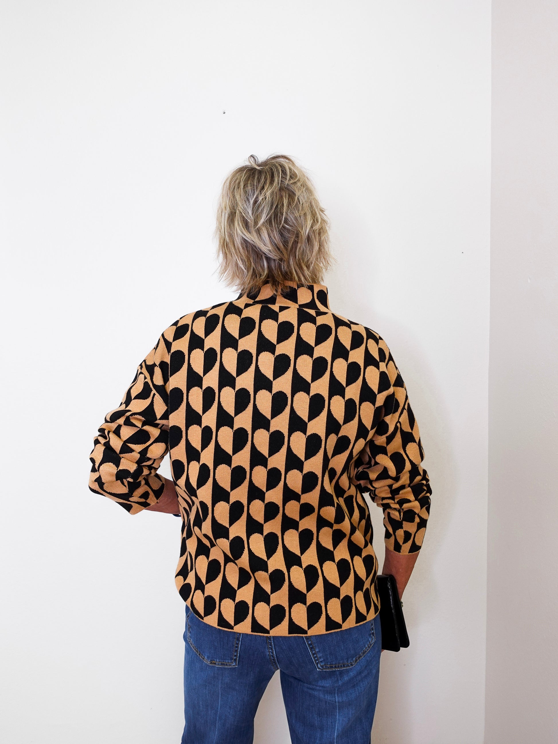 A woman modeling a mock neck heart pattern sweater in front of a white wall. 