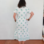 Person wearing a white dress with green patterns standing against a white wall.