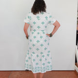 Person wearing a white dress with green patterns standing against a white wall.