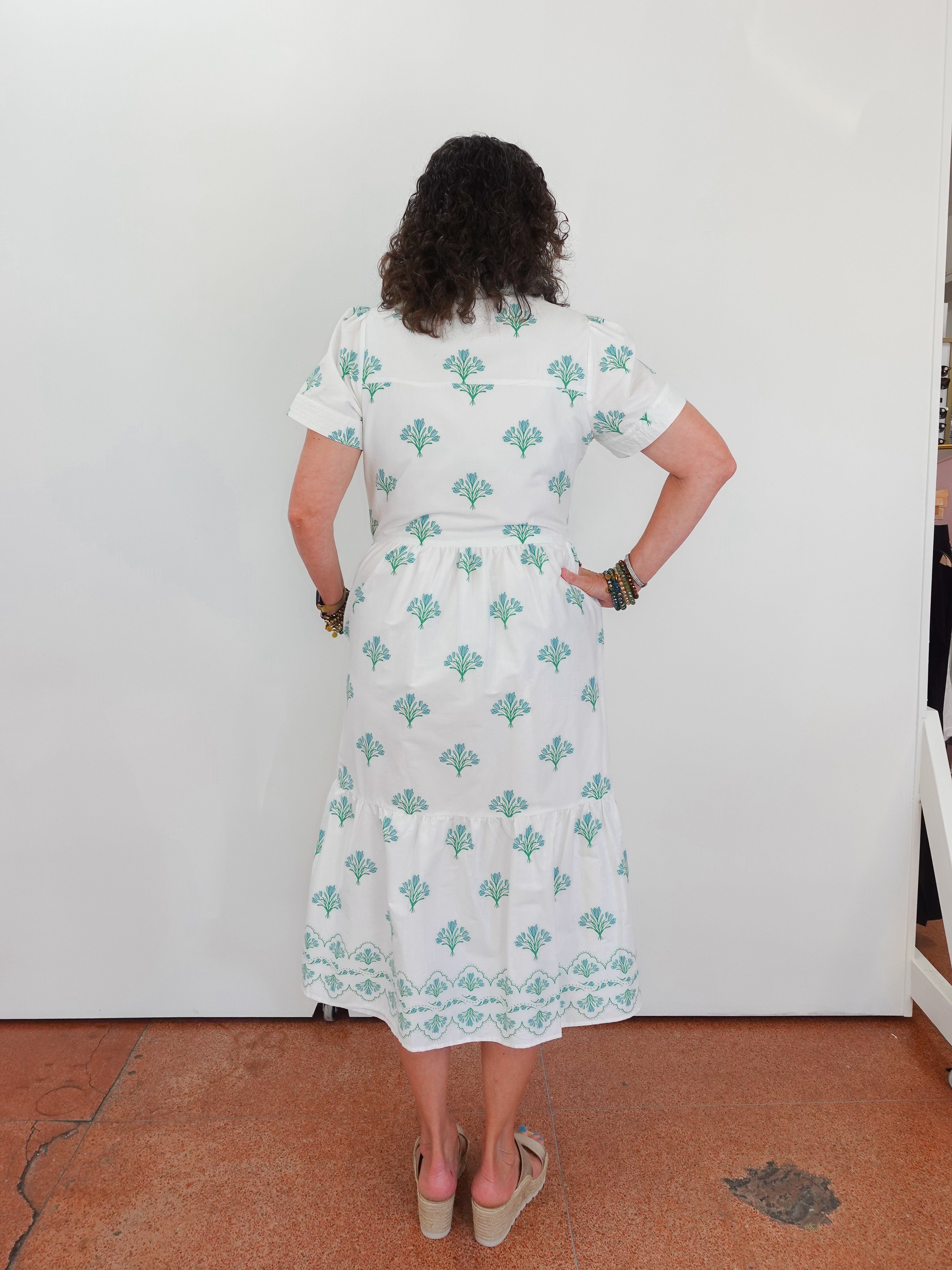Person wearing a white dress with green patterns standing against a white wall.