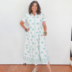 Woman wearing a white dress with green patterns standing against a white wall.