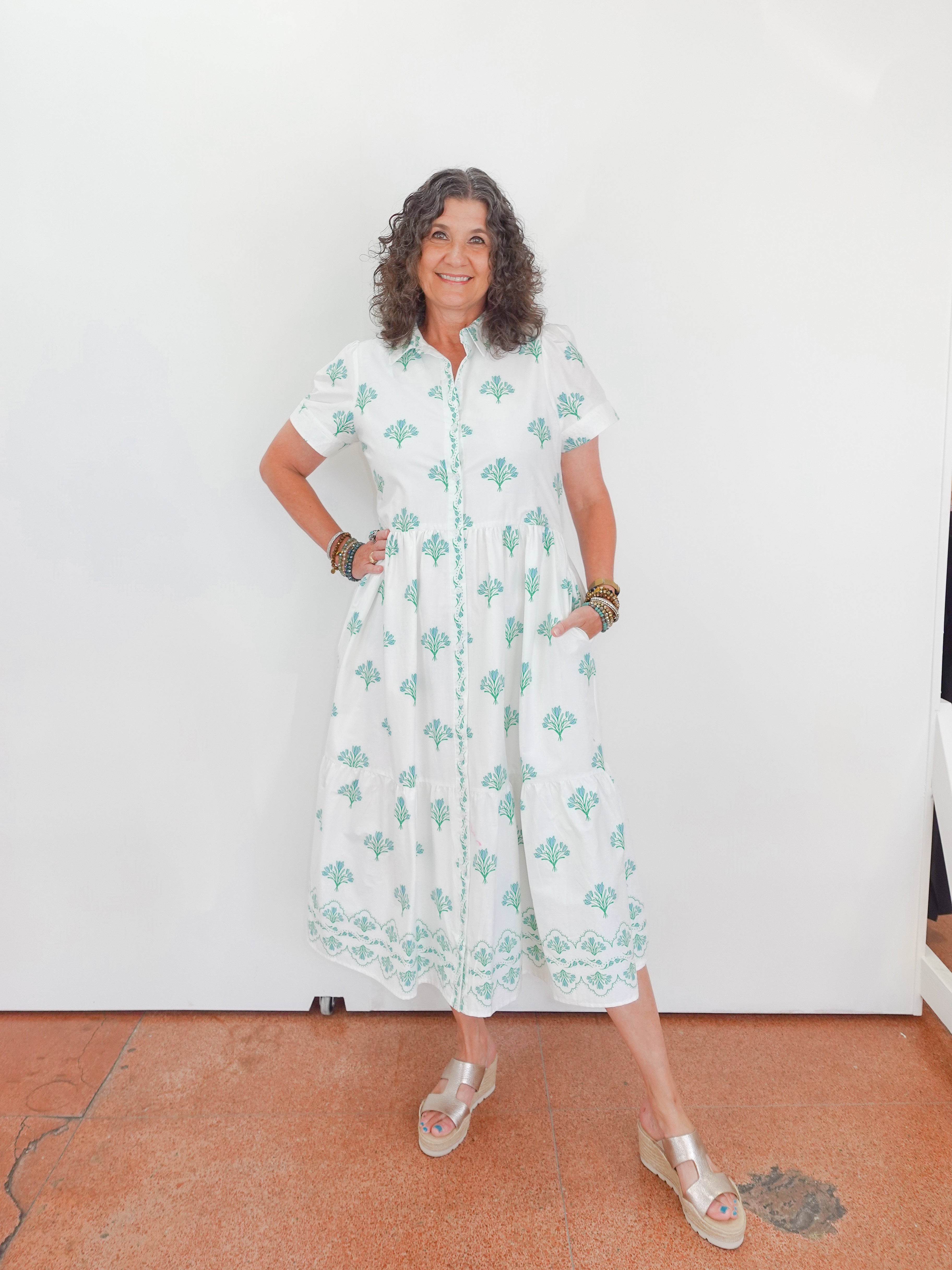 Woman wearing a white dress with green patterns standing against a white wall.