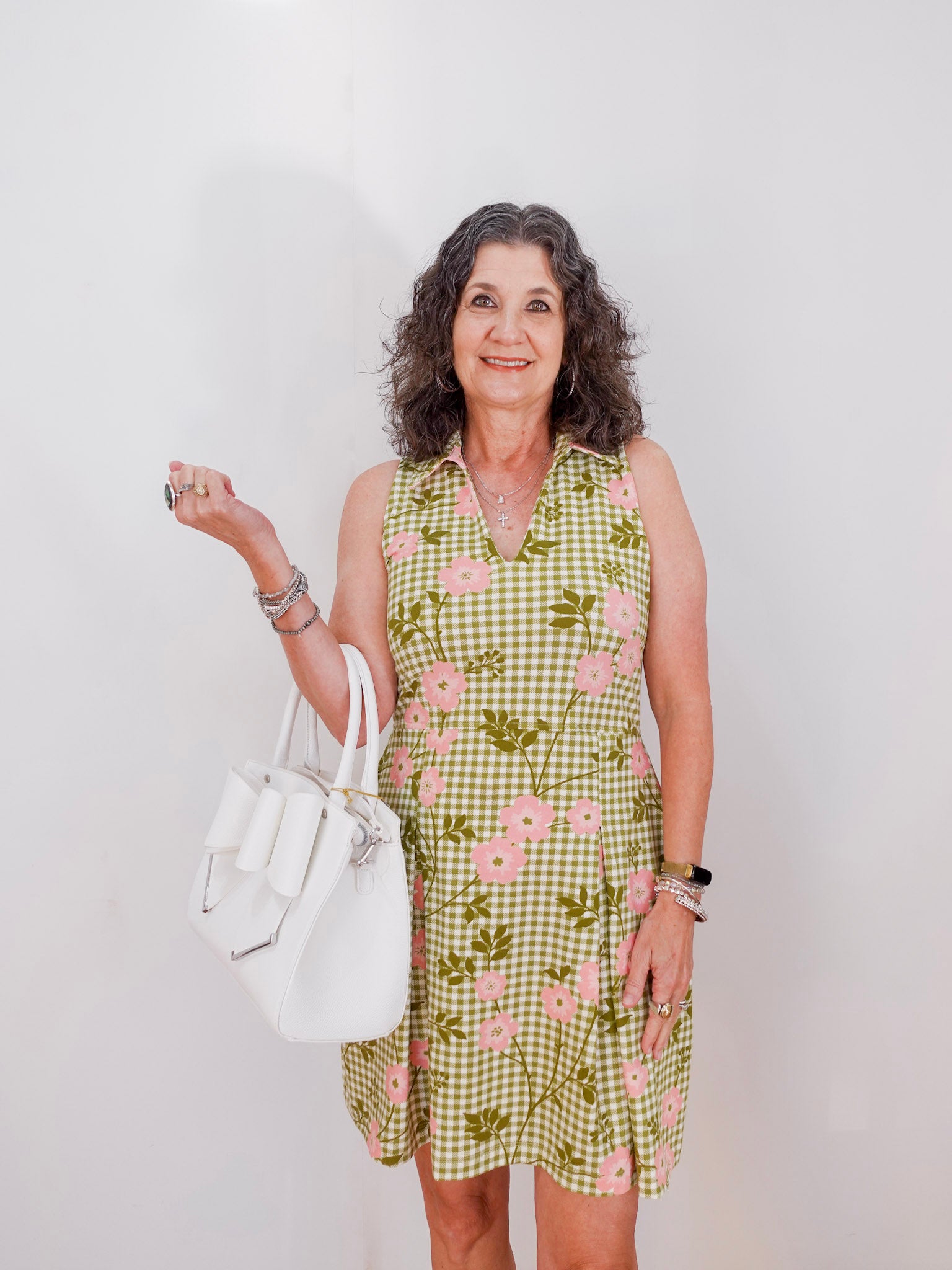 Woman wearing a green checkered dress with floral patterns, holding a white handbag against a plain background.