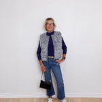 Person wearing a gray vest, navy sweater, blue jeans, and white sneakers against a white wall.