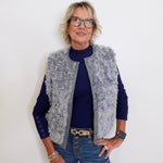 Person wearing a gray fur vest over a blue sweater and jeans against a white background