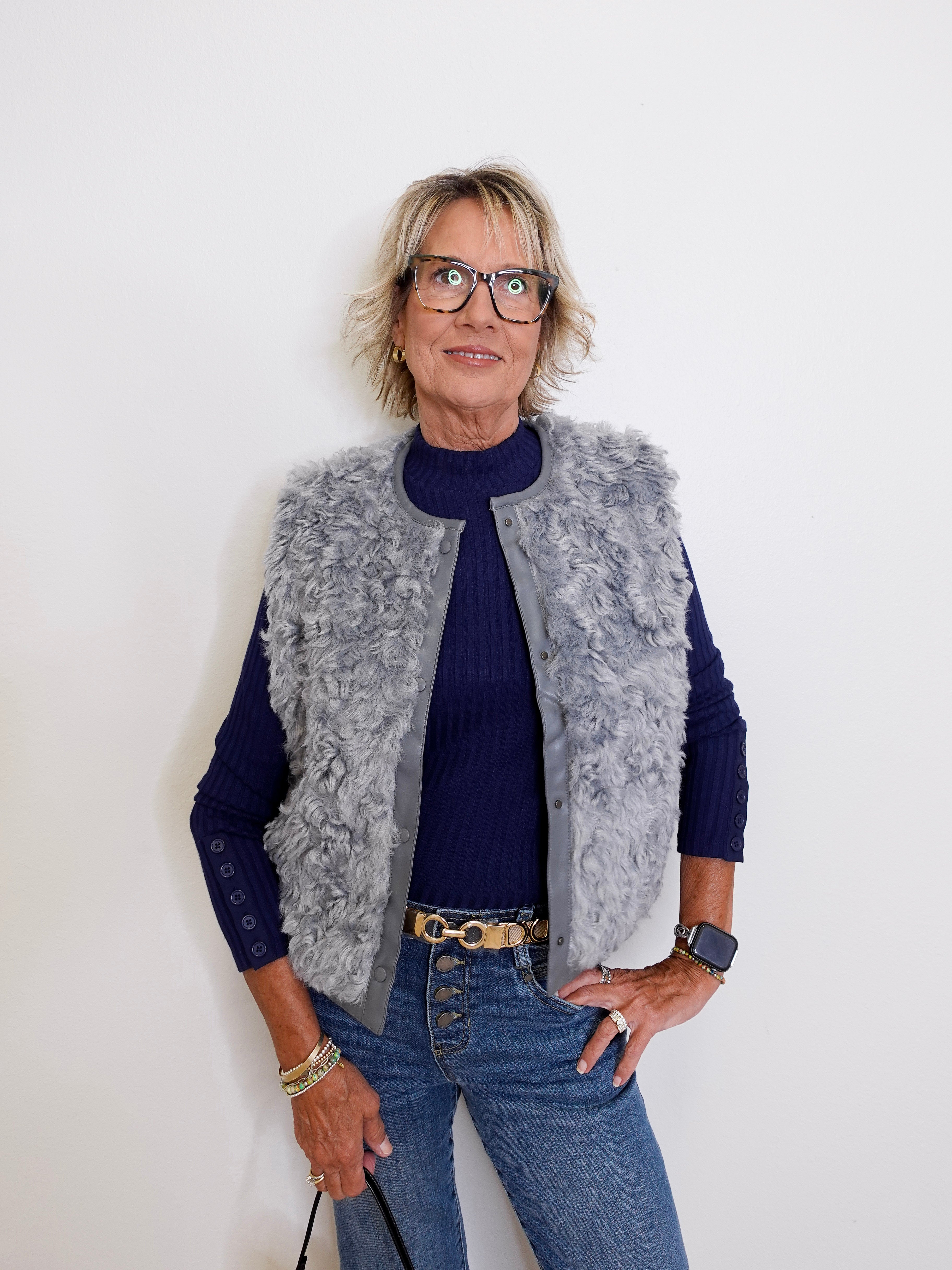 Person wearing a gray fur vest over a blue sweater and jeans against a white background