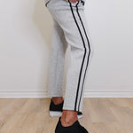 Person wearing gray sweatpants with black stripes and black sneakers on a light wooden floor.