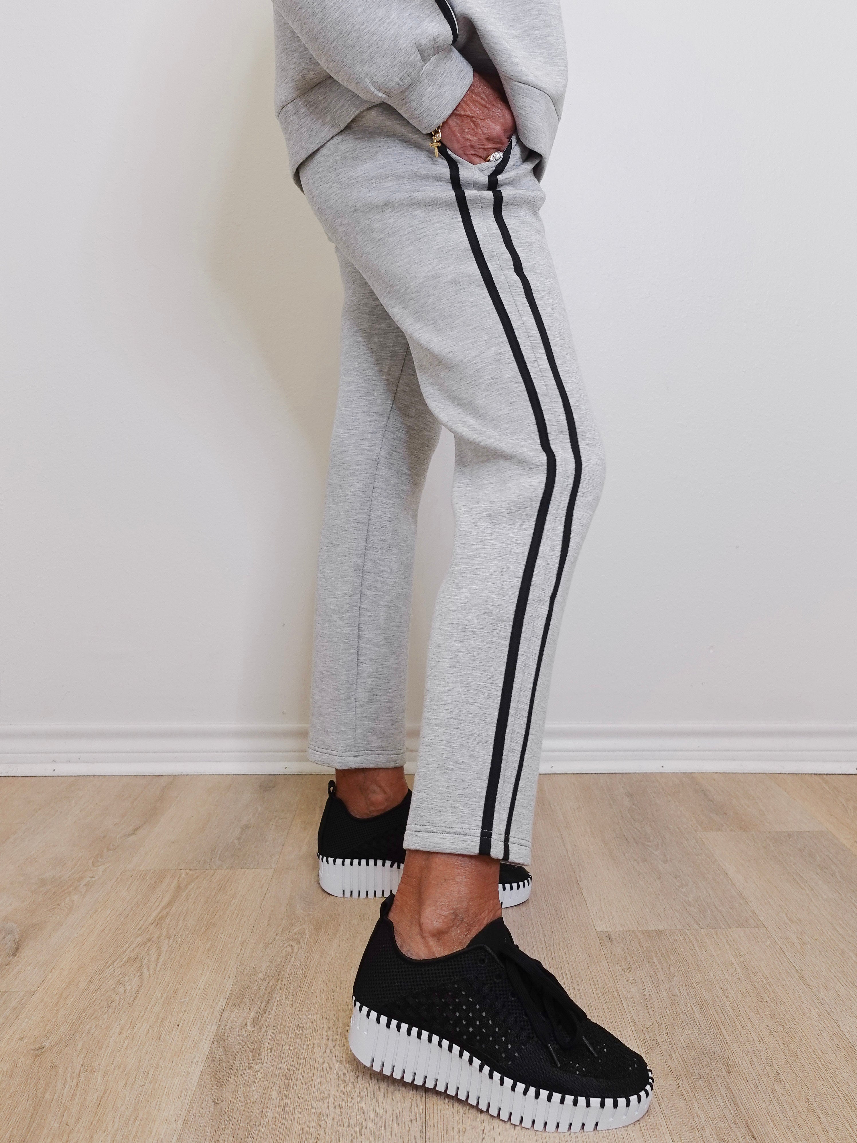 Person wearing gray sweatpants with black stripes and black sneakers on a light wooden floor.